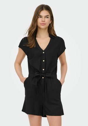 Junge Frau trägt einen schwarzen kurzärmeligen, geknöpften Overall mit Taillengürtel, steht mit den Händen in den Taschen und blickt leicht nach rechts.