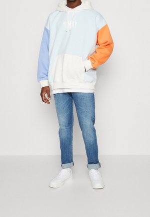 Mikina s kapucí v blokových barvách světle modré, oranžové a krémové, s přední kapsou a bílým logem. Kombinována s modrými džínami a bílými teniskami.