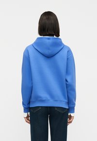 Άτομο με κοντά σκούρα μαλλιά που φοράει μια φωτεινή μπλε μπλούζα με κουκούλα και σκουρόχρωμο τζιν, στέκεται με την πλάτη προς τον θεατή μπροστά σε απλό φόντο.