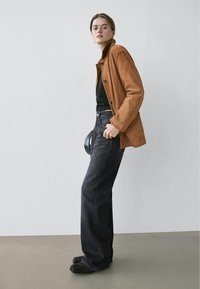 Hnědá semišová bunda s vysokým límcem, černý top a tmavé džínové kalhoty. Oblečení má volný střih a zahrnuje černé boty.