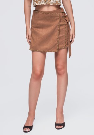Mujer con falda cruzada de ante marrón con tachuelas metálicas y lazo lateral, combinada con un top corto beige y tacones negros abiertos en la punta.