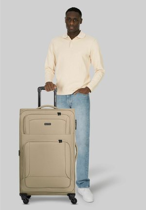 Mann in cremefarbenem Langarmshirt und Jeans steht und hält den Griff eines großen beigen Koffers mit vier Rollen vor grauem Hintergrund.