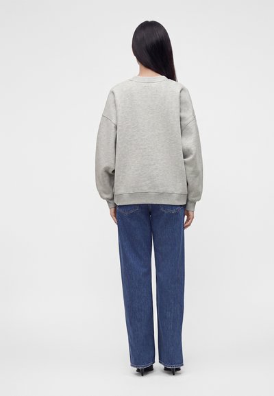 Personne aux longs cheveux noirs, de dos, portant un sweat-shirt gris clair, un jean droit bleu et des chaussures noires pointues.