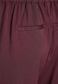Pantalons en tissu bordeaux avec une finition texturée, dotés d'une taille élastique et d'une poche arrière unique avec une fermeture plate.