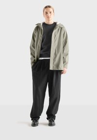 Chaqueta de lluvia verde oliva claro con capucha, cierre de botones a presión y forro interior blanco en contraste, combinada con pantalones negros holgados y zapatos negros.
