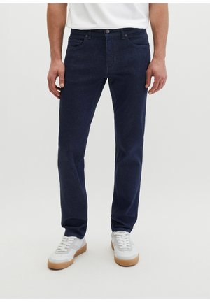 Mann trägt dunkelblaue Jeans mit geradem Bein und weiße Sneakers mit Gummisohlen, steht vor einem einfarbigen hellen Hintergrund.