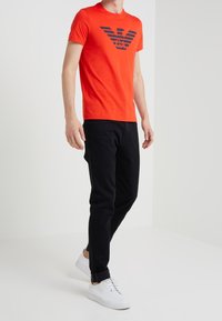Camiseta roja de manga corta con un logo de alas en negro, combinada con pantalones ajustados negros y zapatillas blancas. Tela suave, diseño atlético y casual.
