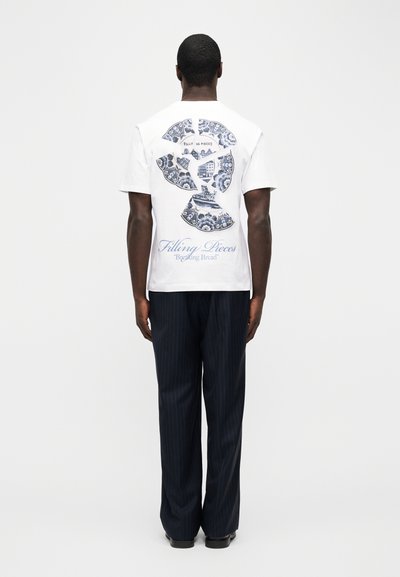 Άνδρας που φοράει λευκό μπλουζάκι με σχέδιο σπασμένου πιάτου και το κείμενο «Filling Pieces 'Breaking Bread'», συνδυασμένο με σκούρο παντελόνι με ρίγες και μαύρα παπούτσια.