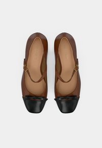 Chaussures Mary Jane marron avec brides ajustables, bouts noirs à capuchon et petits nœuds, présentées côte à côte sur un fond blanc.