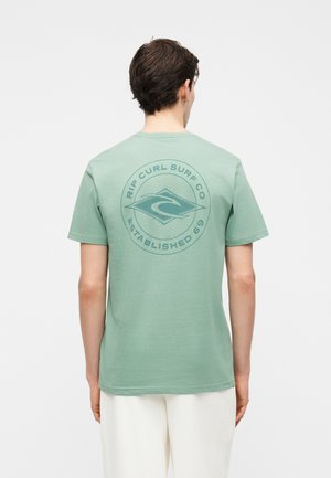 Mann med kort mørkt hår iført en lysegrønn Rip Curl Surf Co t-skjorte og hvite bukser, vist bakfra på ensfarget bakgrunn.