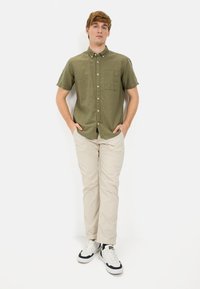 Olijfgroene overhemd met korte mouwen en knopen, met een borstzakje, gecombineerd met crèmekleurige pantalon en witte sneakers met marineblauwe accenten.
