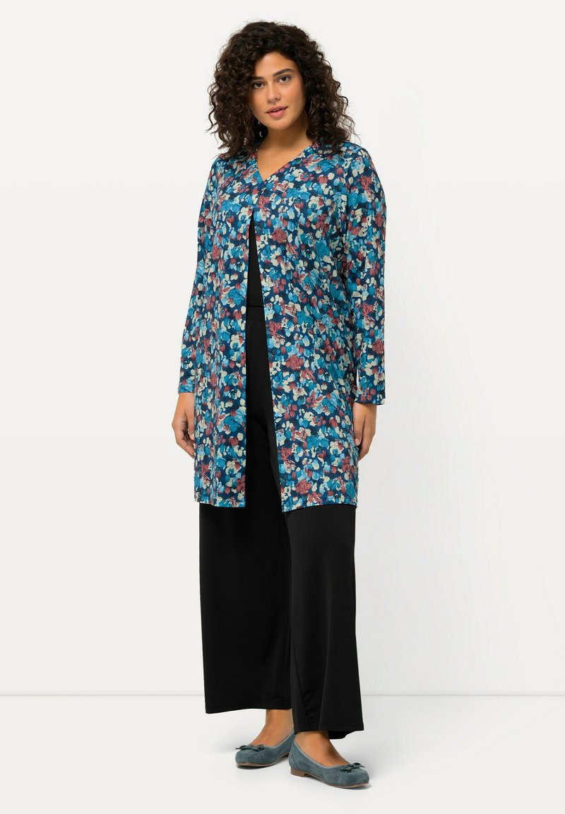 Cardigan largo floral con base azul y patrones rojos y blancos, combinado con pantalones anchos negros y zapatos planos azules. Textura de tela suave.