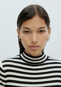 Ein Modell trägt einen schwarz-weißen gestreiften Ripp-Turtleneck. Die Haare sind glatt und glänzend, und silberne Creolen ergänzen das Outfit.