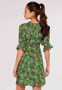 Grünes Blumenkleid mit kurzen Rüschensäumen, das ein Muster aus kleinen weißen, orangefarbenen und rosa Blumen auf einem dunkelgrünen Hintergrund zeigt.