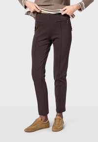Braune maßgeschneiderte Hose aus Stretchstoff, mit Taschen vorne, schmaler Passform und Reißverschlussakzenten. Kombiniert mit hellbraunen Turnschuhen.