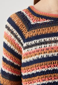 Gros plan d'un pull en crochet avec des rayures horizontales bleu marine, blanc, marron, rose et gris, et un motif ajouré sur l'épaule et le haut de la poitrine d'une personne.