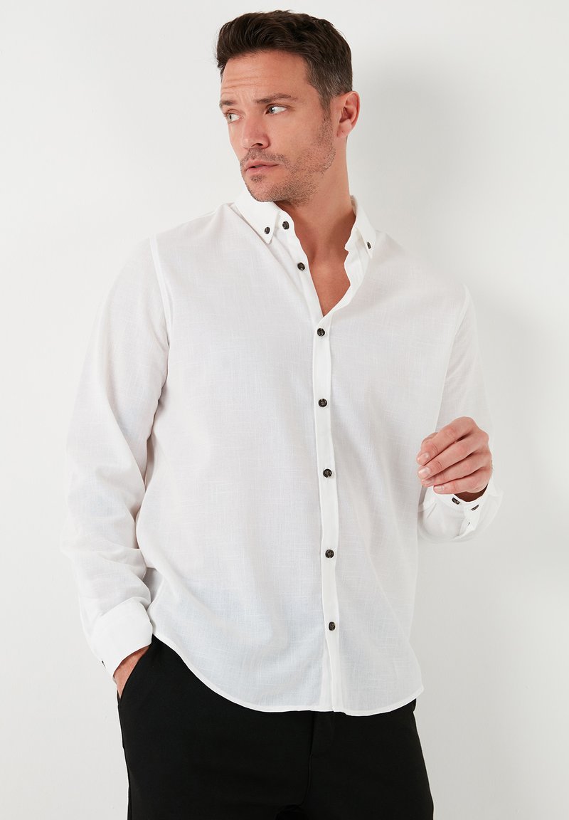Camisa blanca de manga larga con cuello abotonado, botones negros y una tela texturizada. Combinada con pantalones negros, con las manos colocadas de forma casual.