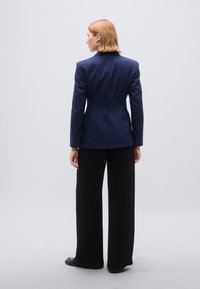 Μπλέιζερ ναυτικού με ρίγες, στενή γραμμή, γιακάδες με σχισμές και μακριά μανίκια, σε συνδυασμό με μαύρο παντελόνι με φαρδιά πόδια. Απλό φόντο.