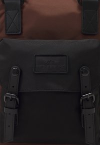 Schwarzer Stoffrucksack mit einem oberen Abschnitt in Braun, ausgestattet mit einer Frontklappe, die zwei schwarze Schnallen und ein Logopatch aufweist. Robuste Träger und Texturen.