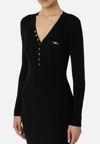 Vestito nero a coste con maniche lunghe, scollo a V profondo e dettagli con bottoni dorati. Presenta una tasca frontale con un accento dorato. Texture liscia.