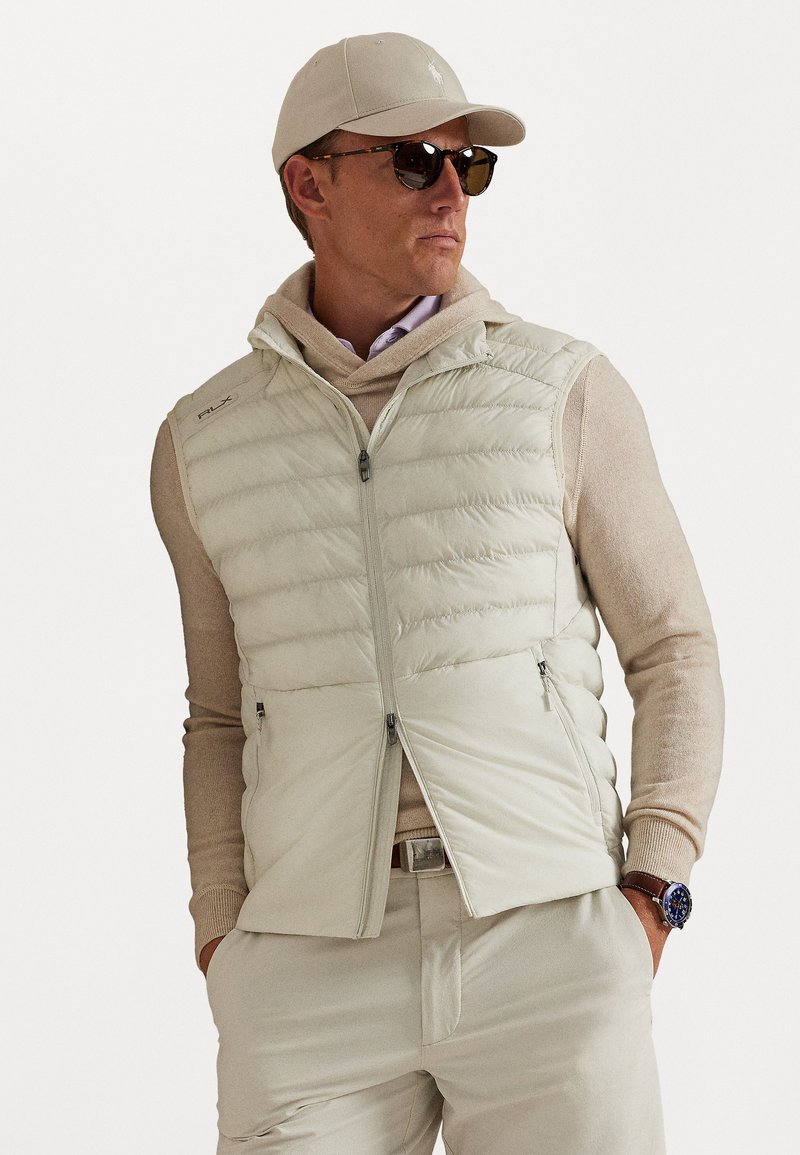 Homme portant une casquette beige, des lunettes de soleil, un gilet matelassé par-dessus un hoodie, un pull à manches longues et un pantalon léger, les mains dans les poches.