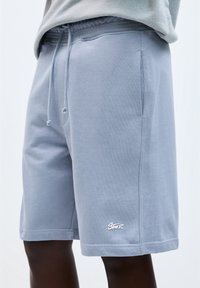 Shorts en sweat léger bleu avec une taille élastique, poches latérales et un petit logo blanc en bas de l'ourlet. Tissu doux et coupe décontractée.