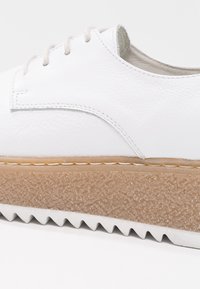 Chaussure en cuir blanc avec une semelle plateforme beige texturée et une bande de roulement en zigzag blanche, dotée de lacets blancs et de détails de couture visibles.