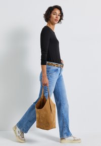 Donna che cammina indossando jeans azzurri chiari, top a maniche lunghe neri, cintura con stampa leopardata, sneakers beige, mentre porta una grande borsa in suede color cammello.