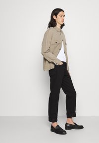 Camisa beige de botones con cuello y bolsillos de solapa, llevada sobre una camiseta blanca. Pantalones negros entallados y mocasines negros, de pie sobre un fondo claro.