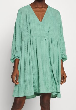 Žena v volných, texturovaných mentolově zelených šatech s výstřihem do V a dlouhými nabíranými rukávy, stojící před jednoduchým bílým pozadím.