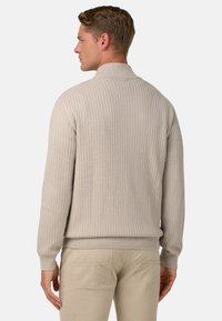 Szary sweter prążkowany z wysokim kołnierzem, o strukturalnej tkaninie i dopasowanych rękawach. Prezentowany na modelce odwróconej w stronę, zestawiony z jasnymi spodniami.