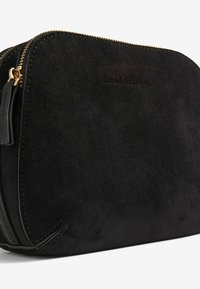 Borsa in suede nera con una texture liscia, zip dorata e branding impresso. Forma leggermente arrotondata, con un accento in pelle laterale.