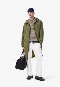 Giacca lunga verde oliva, maglione multicolore, pantaloni bianchi, borsa nera e scarpe scure. Il cappello è blu navy; la borsa ha doppie maniglie.