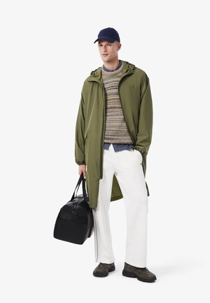 Abrigo largo en verde oliva, suéter multicolor, pantalones blancos, bolso negro y zapatos oscuros. El sombrero es de color azul marino; el bolso tiene dos asas.