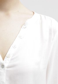 Vit blus med V-ringning och små knappar framtill. Tyget verkar lätt och mjukt med en subtil glans.