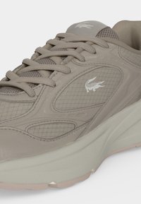 Baskets beige avec des empiècements en mesh et synthétiques, lacets beiges, logo crocodile blanc sur le côté, et semelle amortie.