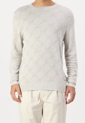 Mężczyzna ubrany w jasnobeżowy sweter z wzorem i długim rękawem oraz dopasowane jasnobeżowe spodnie, stojący na tle jednolitej białej ściany.