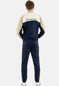 Marineblaue und beige Trainingsanzug aus glattem Stoff. Besitzt eine zipfelbare Jacke mit Logo auf dem Rücken und einem elastischen Bündchen. Bequeme Hosen mit seitlichen Streifen.