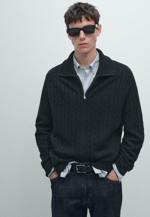 Maglione nero a trecce con colletto alto, dotato di una zip frontale. Indossato sopra una camicia a righe azzurre e jeans scuri, abbinato a occhiali da sole.