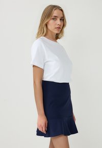Jonge vrouw met blond haar, gekleed in een wit shirt met korte mouwen en een marineblauwe wijde rok, staande tegen een effen lichte achtergrond.