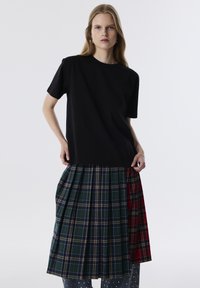 Zwarte korte mouwen t-shirt gecombineerd met een geplooide geruite rok met groene, blauwe en rode patronen, wat een gelaagd uiterlijk creëert.