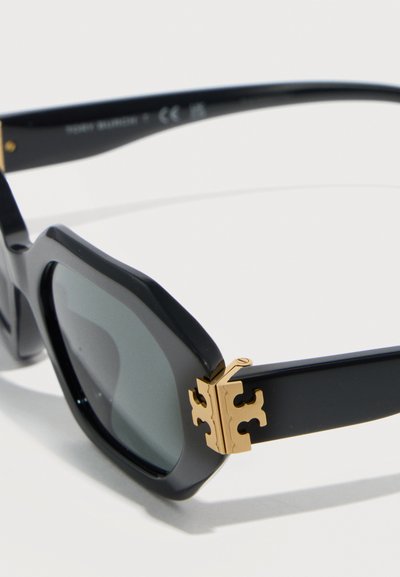 Lunettes de soleil noires surdimensionnées avec des verres foncés et un détail de charnière dorée sur les branches du cadre, sur fond blanc.