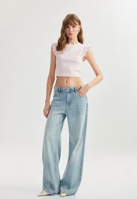 Lichtroze geribbelde crop top met ruffle mouwen, gecombineerd met wijde lichtblauwe denim jeans. Crèmekleurige schoenen. Minimalistische setting.