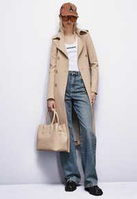 Bežový trenčkot so zlatými gombíkmi, biely top bez rukávov, svetlo modré džínsy, béžová kabelka, hnedý klobúk a veľké slnečné okuliare, čierne topánky.