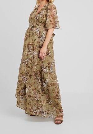 Vrouw in een groene bloemenwikkeljurk met zwierige mouwen, staand tegen een effen lichte achtergrond die de volledige lengte en het vloeien van de jurk toont.