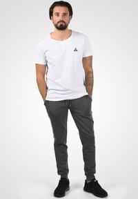 Vit t-shirt med en liten svart triangel-logotyp, tillsammans med grå joggbukser och svarta sportskor. Bakgrunden är neutral och minimalistisk.