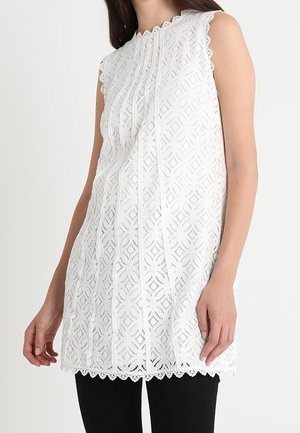Tunique blanche sans manches en dentelle avec des motifs géométriques portée sur un pantalon noir par une femme aux cheveux foncés.