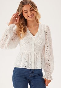 Blusa bianca con motivo ad occhio, scollatura a V e chiusura con bottoni. Presenta maniche lunghe a sbuffo con polsini arricciati. Forma fluida e morbida.
