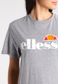 Siva pamučna majica s bijelim "ellesse" logotipom i polukružnim crvenim i narančastim akcentom. Kratki rukavi i okrugli izrez.