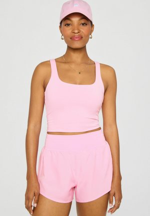Woman wearing light pink athletic tank top, matching high-waisted shorts, and a pink cap, standing against a plain light background.